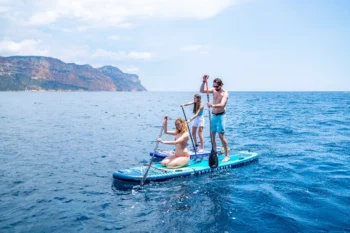 A family of three enjoys a sunny day on the open sea aboard the Aqua Marina SUPER TRIP 12’6″ inflatable stand-up paddleboard from the Family Series: one person kneels at the front paddling, while two others stand toward the rear, all smiling as they glide across deep blue water with dramatic coastal cliffs in the background.